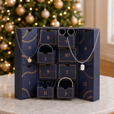 A Christmas advent calendar for jewelry, featuring 12 numbered drawers in dark navy blue with gold accents, displayed on a marble table in front of a blurred Christmas tree.