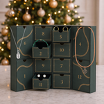 A dark green Christmas advent box with twelve numbered drawers, styled with jewelry including necklaces, earrings, and rings, set on a marble surface with a blurred Christmas tree in the background.