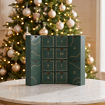 A dark green Christmas advent calendar with twelve numbered drawers and gold decorative details, displayed on a marble table in front of a softly blurred decorated Christmas tree.