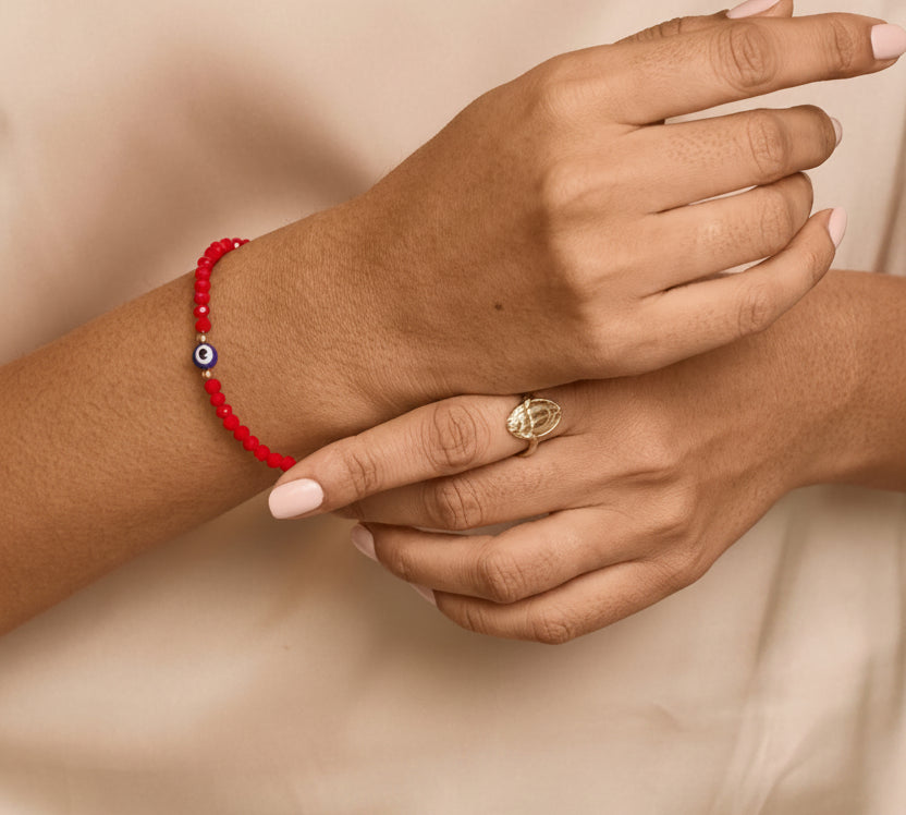 A close-up of a person's hands and arms wearing a red beaded bracelet with a small blue and white evil eye charm, paired with a gold ring, against a soft beige satin top.