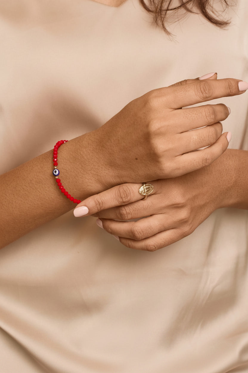 A close-up of a person's hands and arms wearing a red beaded bracelet with a small blue and white evil eye charm, paired with a gold ring, against a soft beige satin top.